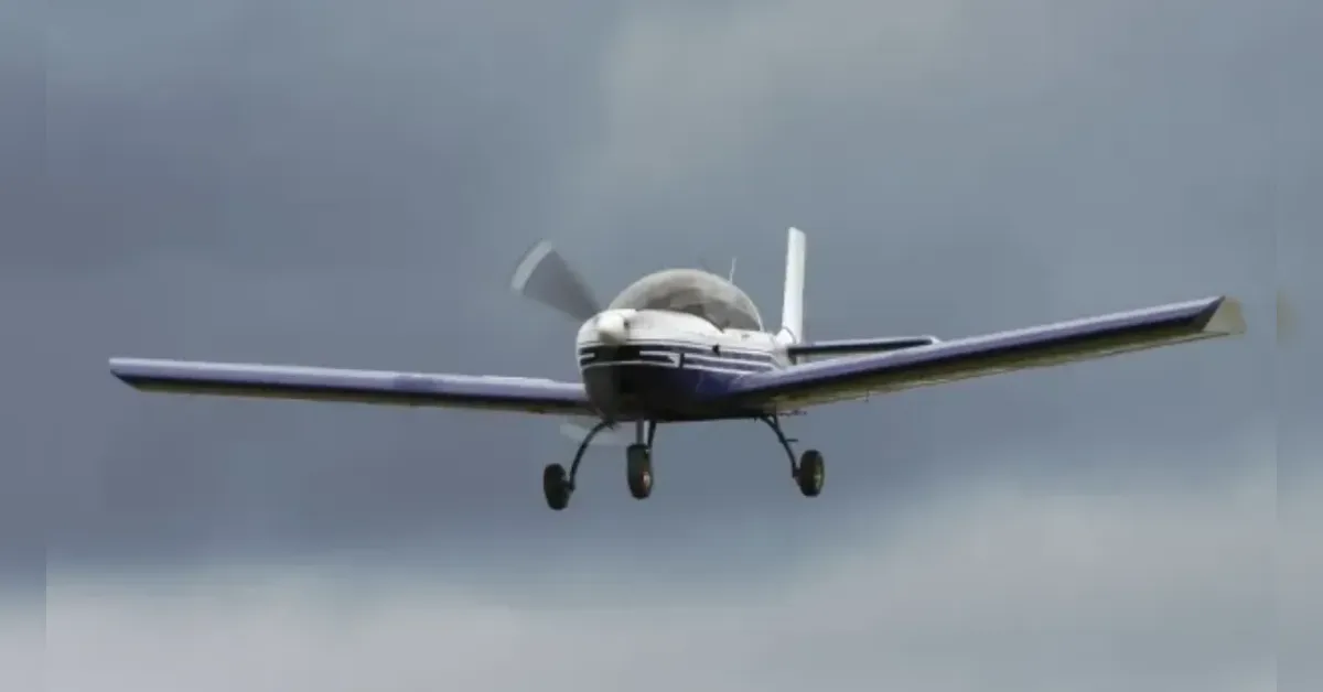 Front view of a small propeller plane in flight toward the camera against a cloudy sky, landing gear down.