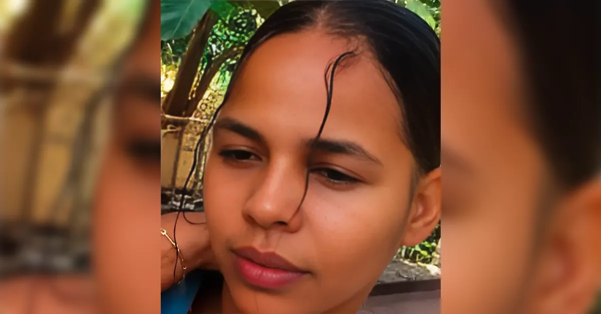 Close-up of a young woman with dark hair resting her chin on her hand, outdoors with greenery in the background.
