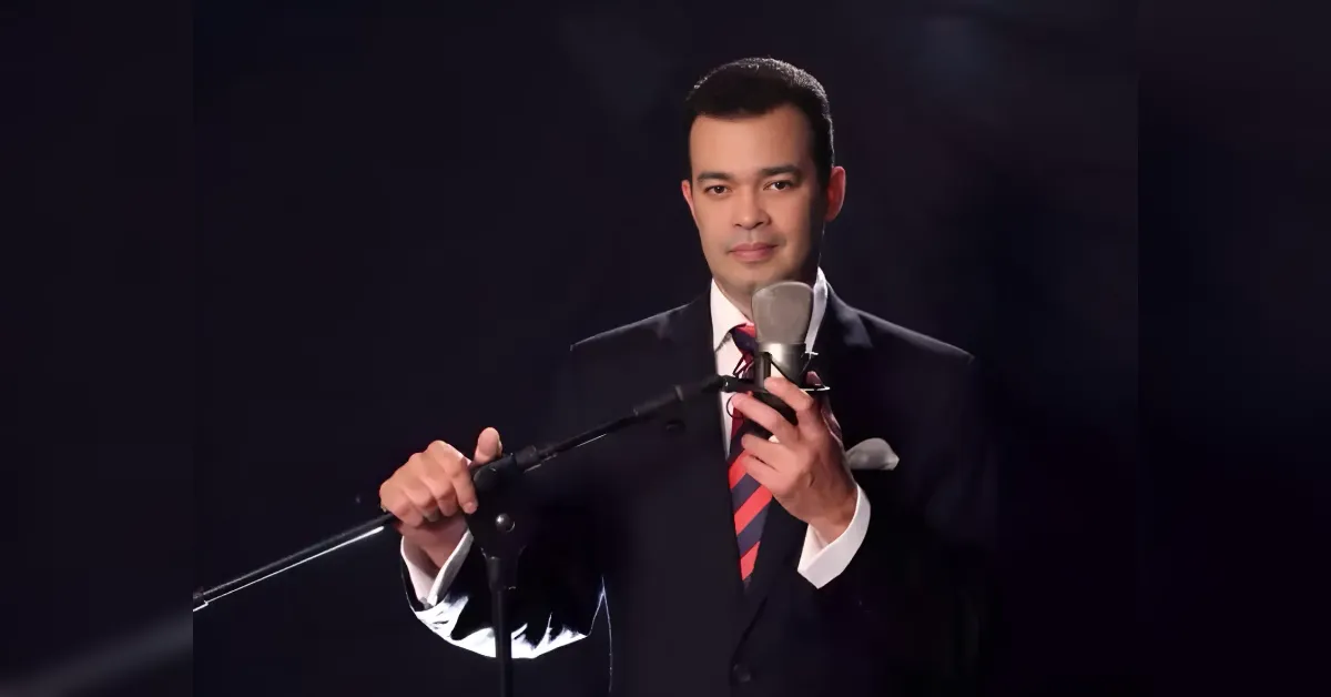 Man in a dark suit and red striped tie speaks into a microphone on a stand against a dark background.