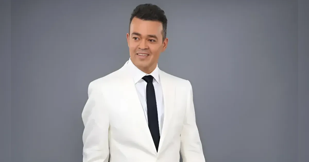 Man in a white suit with a black tie smiles in a studio with a gray background.