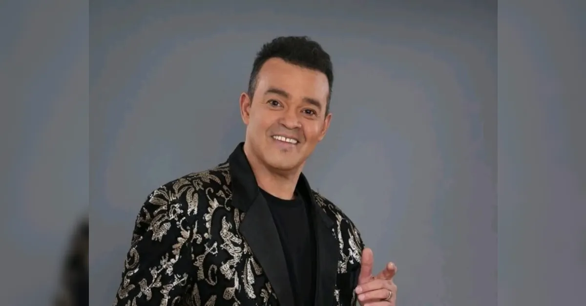 Smiling man in a black shirt and gold-embroidered blazer posing against a gray backdrop.