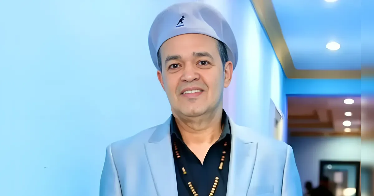 Smiling man in a light blue blazer, black shirt, beaded necklace, and gray beret in a blue-lit hallway.