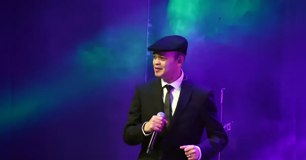 Male singer in a black suit and flat cap performing on stage, holding a microphone with purple-green lighting behind him.