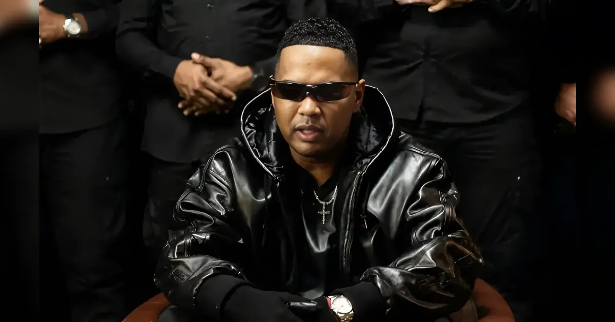 Man in a black leather jacket and sunglasses sits in front of a group of people wearing black, with a visible cross necklace.