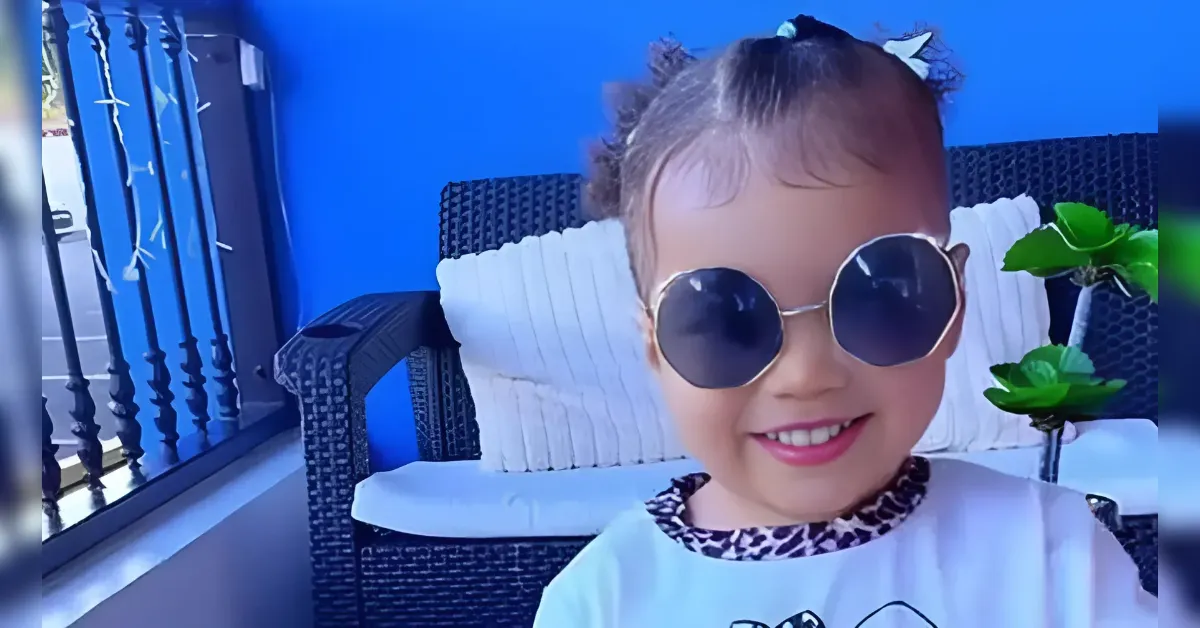 Smiling young child wearing round sunglasses on a balcony with a blue wall and wicker chair behind her.
