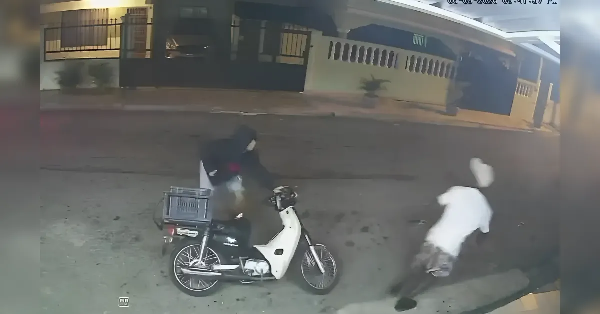 A person in dark clothing sits on a white scooter with a rear cargo box on a dim street at night, while another person in white runs nearby.