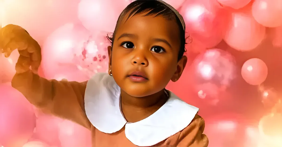 Toddler girl in a brown dress with a white collar, looking at the camera against a pink balloon backdrop.