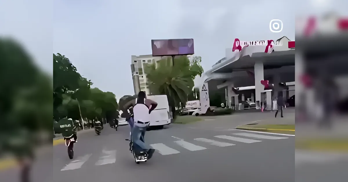 Person on a scooter performing a wheelie in the middle of a crosswalk, other riders nearby on a city street with palm trees and a canopy structure in the background.