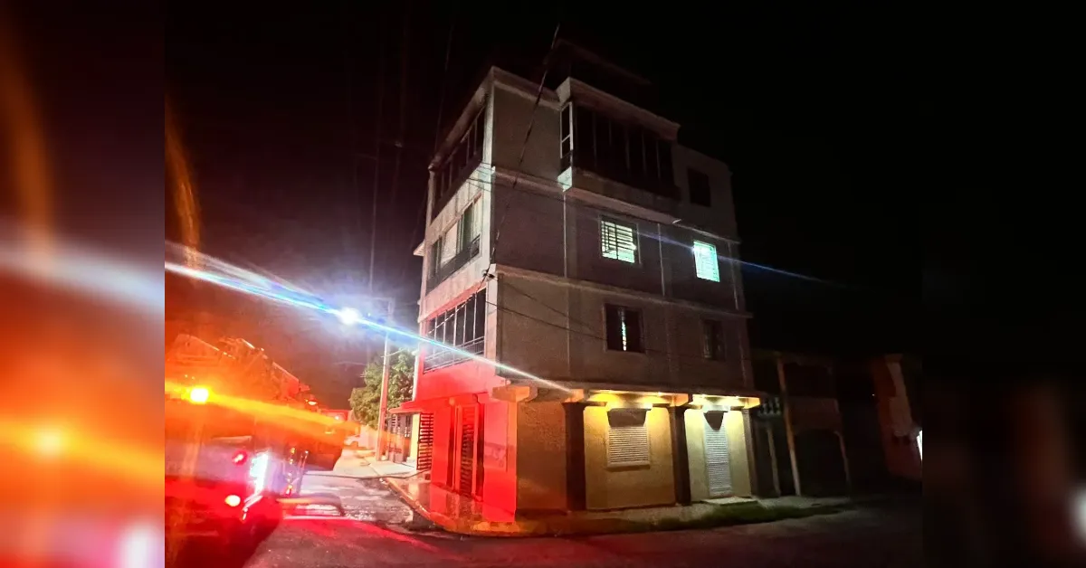 Nighttime street scene with a three-story building and red emergency lights from a fire truck on the left; wet street and illuminated storefronts.