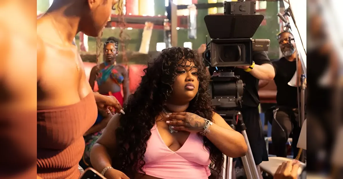 Woman with long curly hair in a pink top sits in front of a large video camera on a film set, with crew members and equipment around her.