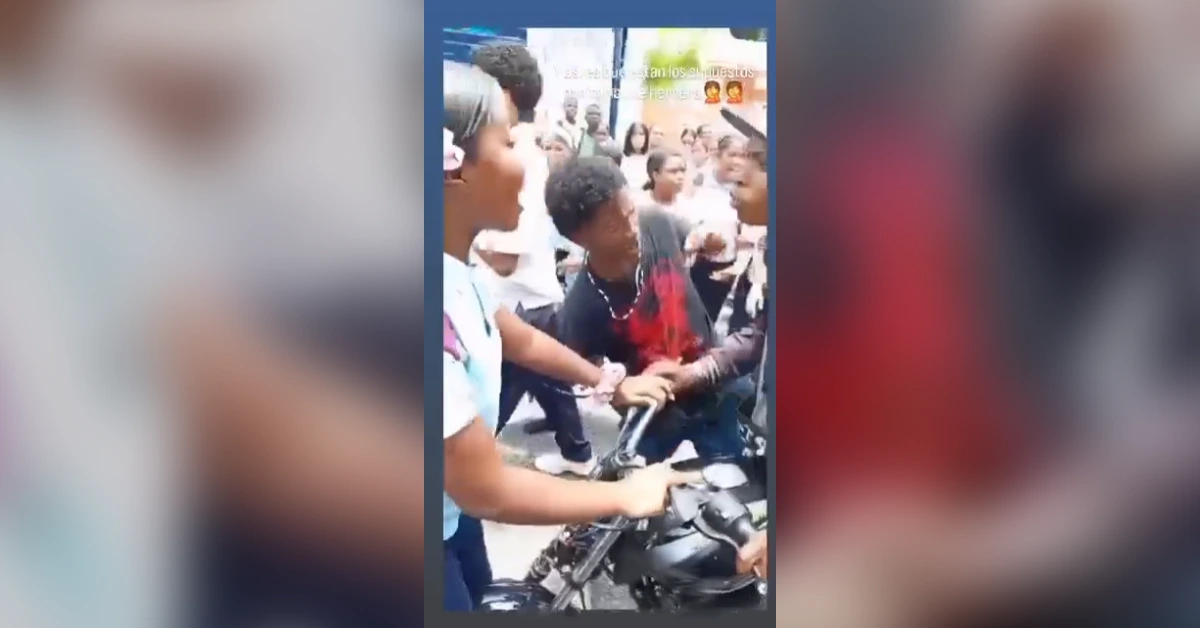 A young man wearing a red-and-black shirt sits on a motorcycle while a crowd crowds around him, grabbing his arms and shirt in a tense street scene.