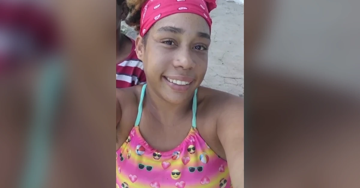 Smiling girl wearing a pink bandana and a colorful emoji-pattern swimsuit, taking a selfie on the beach.