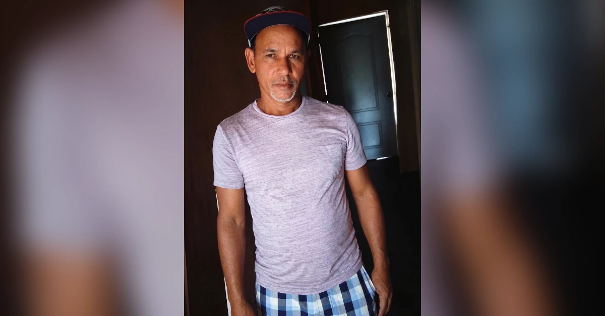 Man in a light gray T-shirt and blue plaid shorts stands indoors, facing the camera with a cap on.