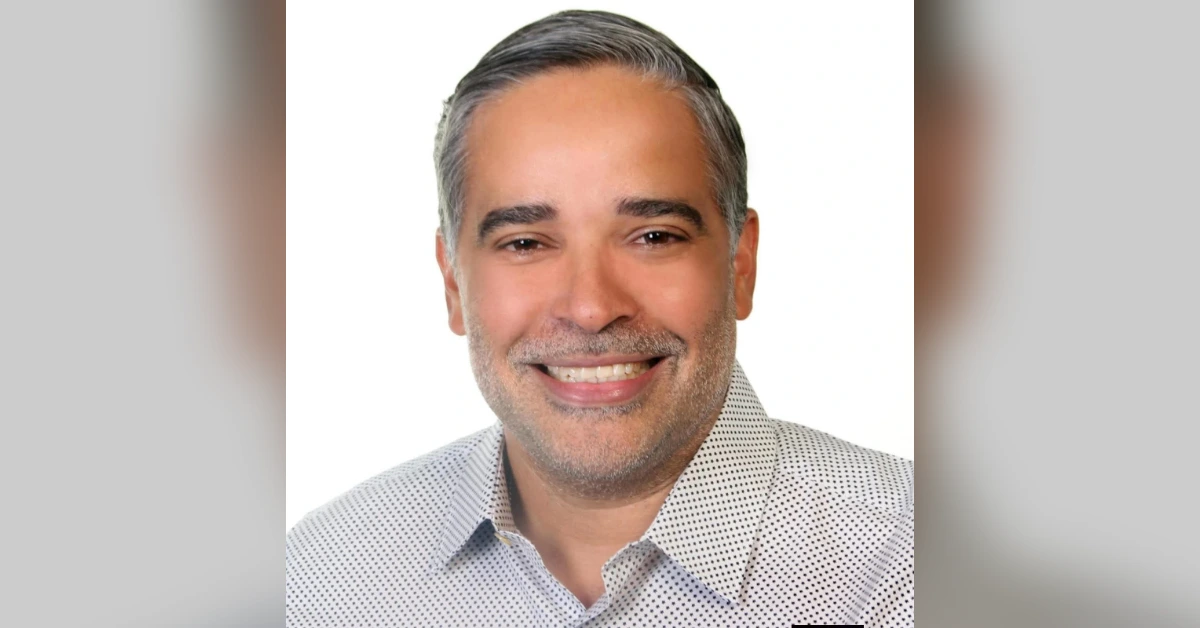 Smiling man with gray hair and stubble wearing a light dotted shirt against a white background, portrait style.
