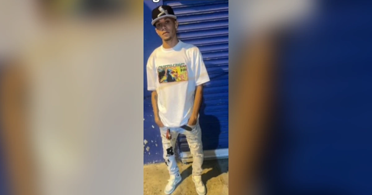 Young man wearing a white graphic T-shirt, ripped light jeans, sneakers, and a cap, standing in front of a blue corrugated wall.