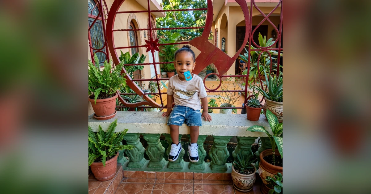 Young child with a blue pacifier sits on a concrete railing amid potted plants, with a decorative red gate behind him.