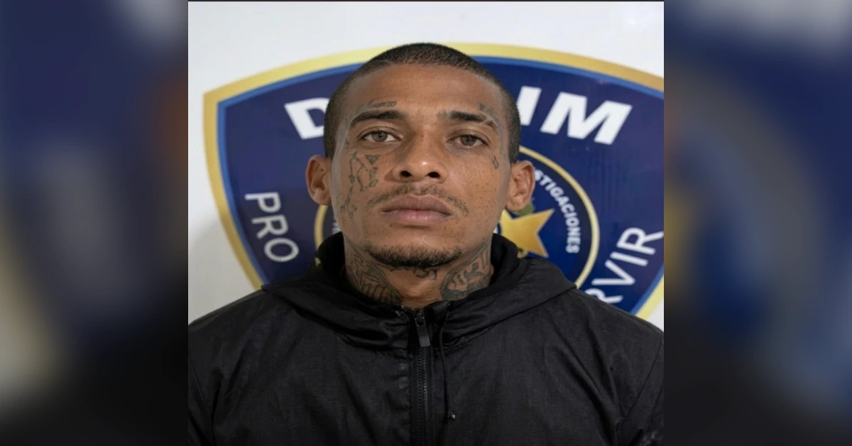 Man with facial tattoos wearing a black zip-up jacket, standing in front of a police emblem backdrop.