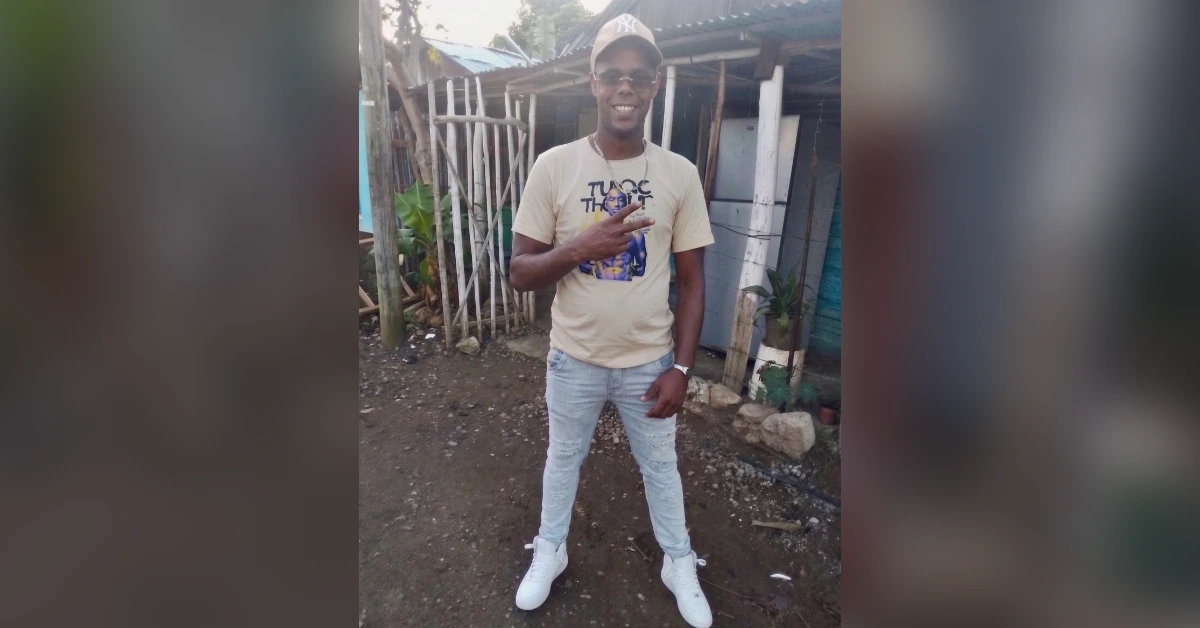 Man wearing a beige graphic T-shirt, light jeans, white sneakers, and a cap, making a peace sign outdoors in a dirt yard.