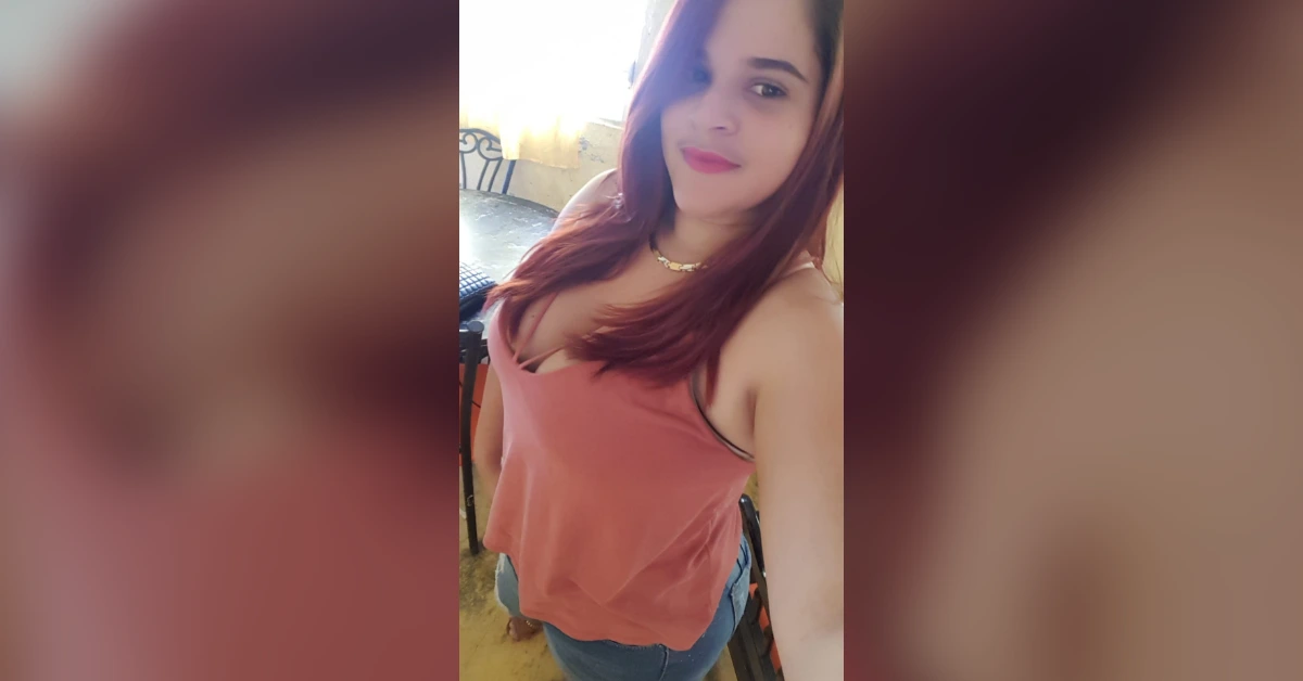 Self-portrait of a woman taking a selfie indoors, wearing a pink sleeveless top and blue jeans, smiling at the camera in soft light