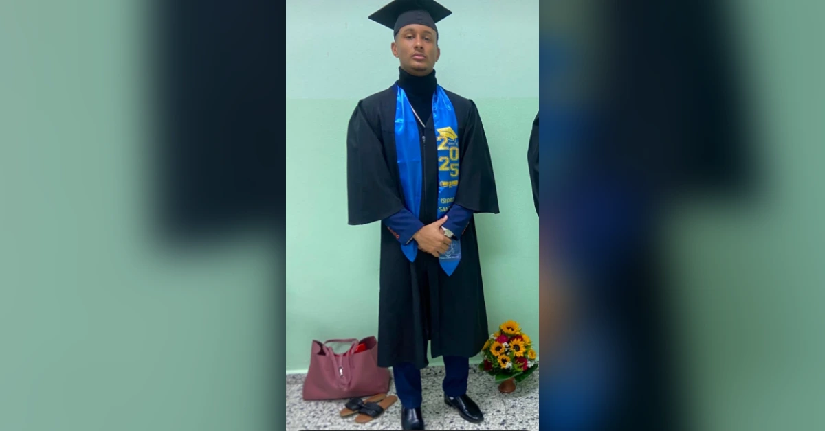 Graduate in a black cap and gown with a blue stole, standing with hands clasped in front of a light green wall; flowers and a purse on the floor nearby.