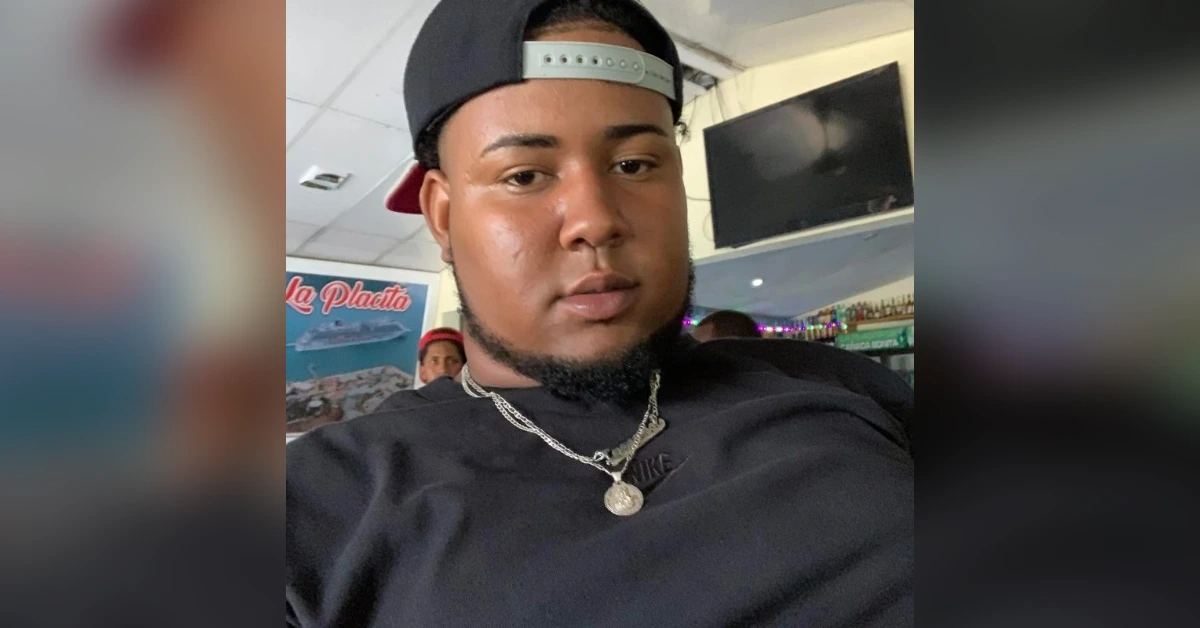 Selfie of a young man wearing a backwards cap and black shirt, with a silver chain and pendant, in a casual indoor setting.