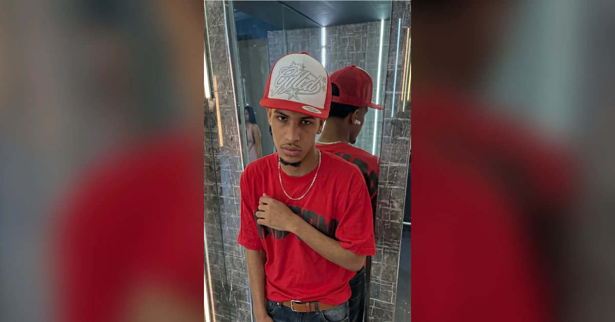 Young man in a red T-shirt and white-red cap poses in a marble-tiled bathroom, hand on chest. Startled look toward camera.