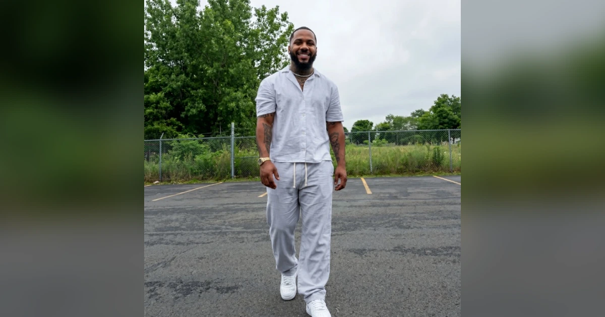 Smiling man with a beard in a light gray loungewear set walking in an empty parking lot wearing white sneakers and a gold watch.
