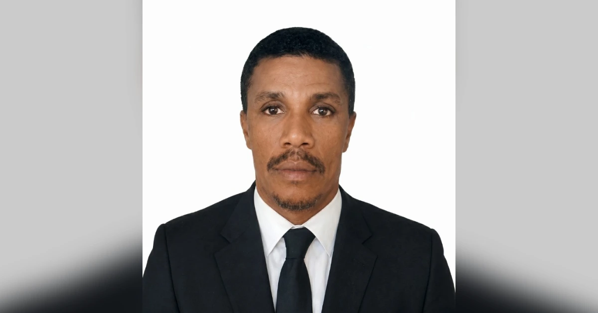 Portrait of a man in a black suit and tie against a white background, looking at the camera with a neutral expression