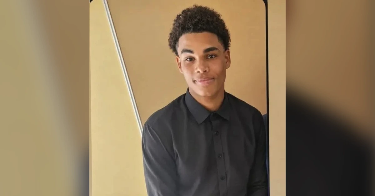 Portrait of a young man with short curly hair wearing a black button-down shirt, standing against a tan wall indoors.