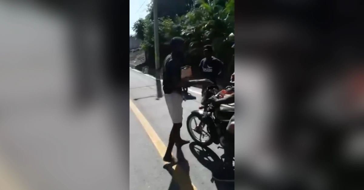 Man in a dark hoodie and white shorts hands something to a person on a motorcycle on a sunny street.