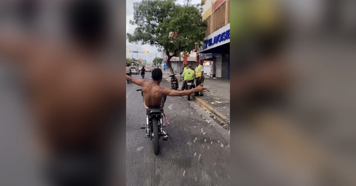 Shirtless man on a motorcycle rides down a city street with arms outstretched toward police near the curb.