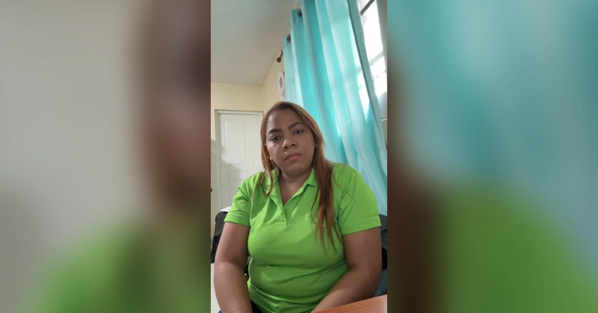 Woman with long hair in a bright green polo shirt sits at a desk in a room with turquoise curtains behind her.