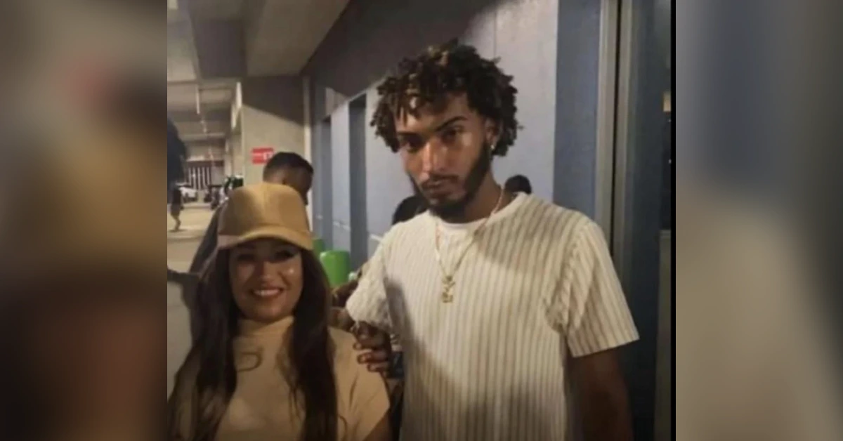 Young man with curly hair in a light striped shirt stands beside a smiling woman in a tan outfit and hat in a hallway.