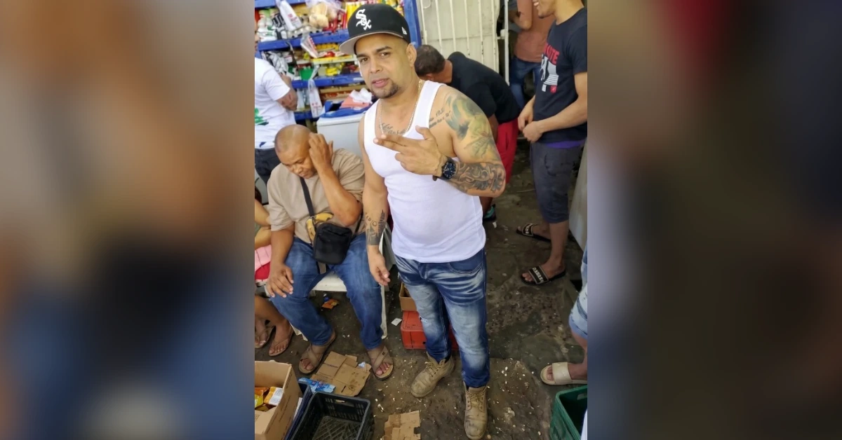 Central man with tattoos in a white tank top and cap poses for the camera in a crowded market stall.