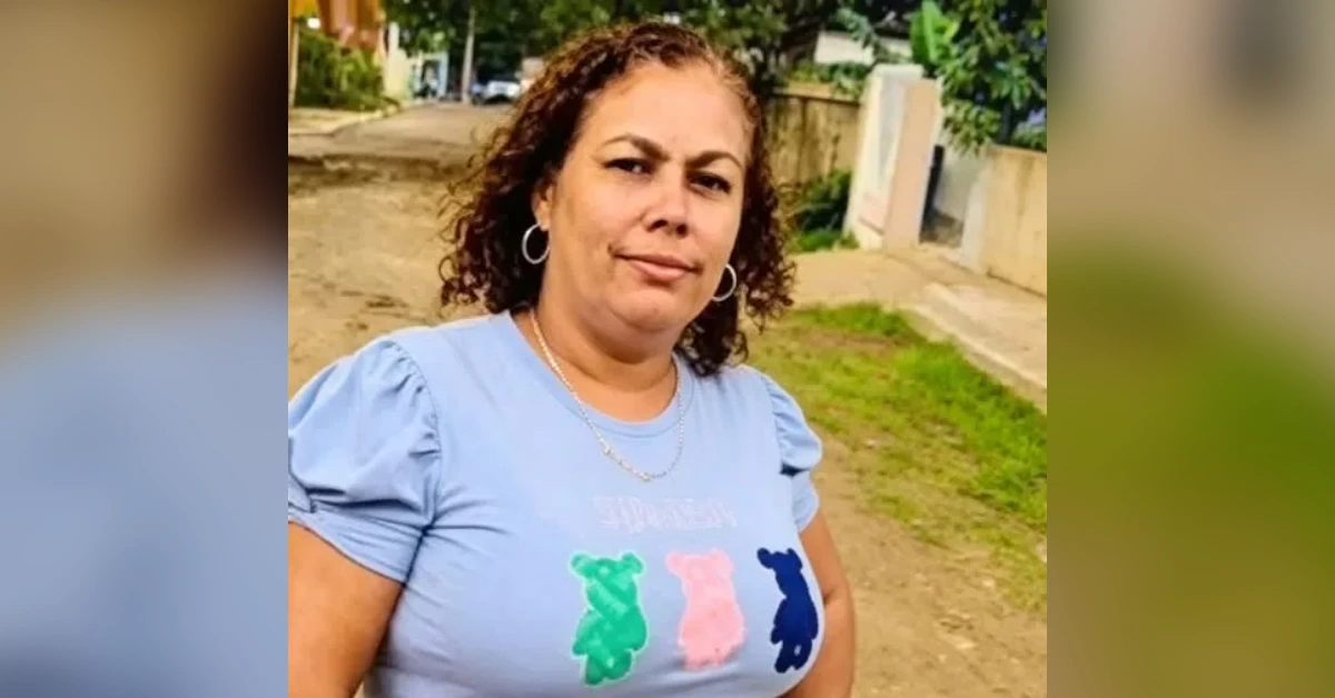Outdoor portrait of a woman with curly hair wearing a light blue shirt with colorful bear silhouettes, standing on a sunny street path in a residential area.