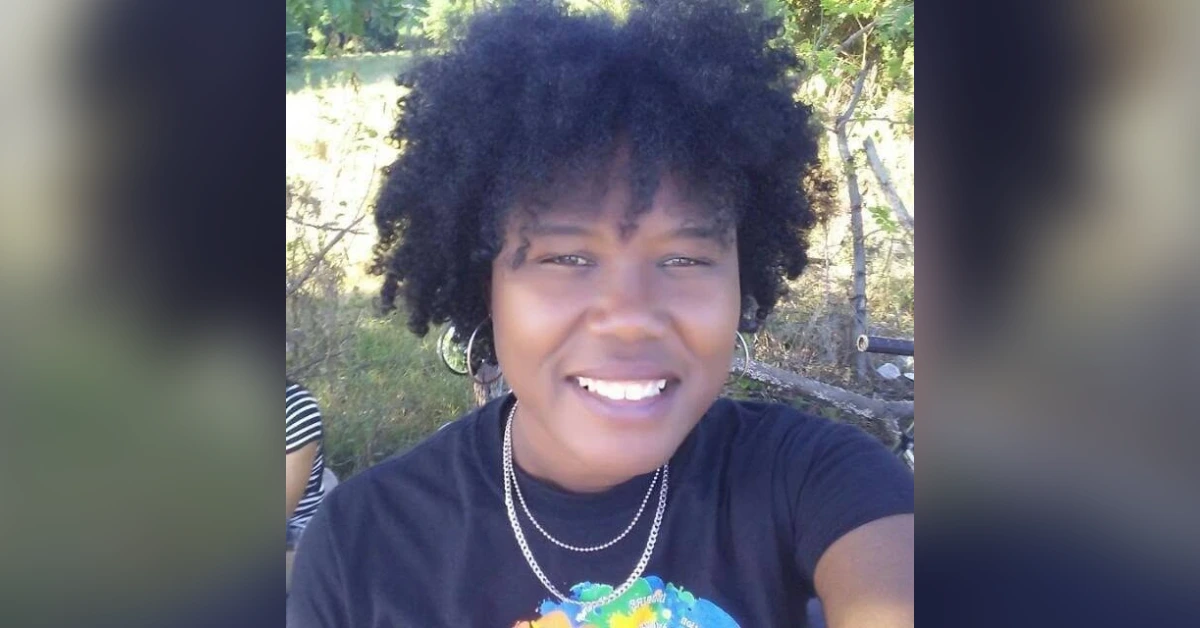 Smiling woman with natural curly hair wearing a black shirt outdoors, trees in the background and hoop earrings.