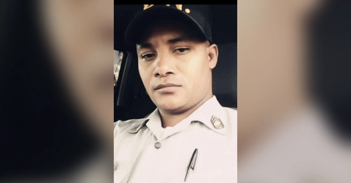 Man in a light-colored uniform shirt and cap sits inside a vehicle, looking at the camera with a neutral expression.