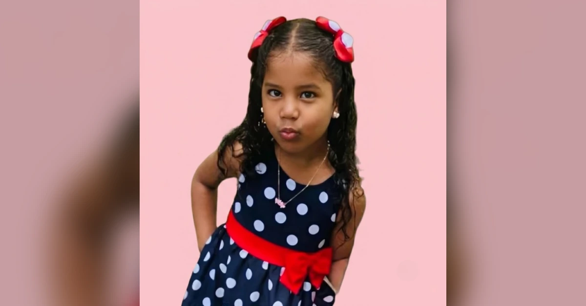 Young girl in a navy polka-dot dress with a red sash posing with a pout against a pink background.