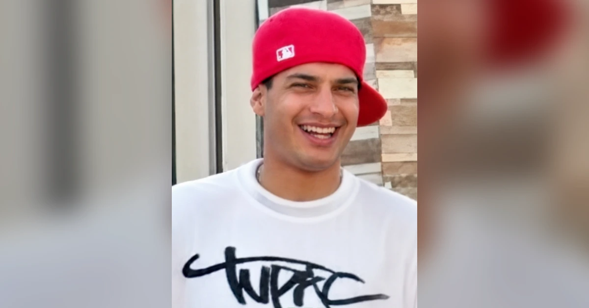 Smiling man wearing a red backward baseball cap and white graphic tee outside a building.
