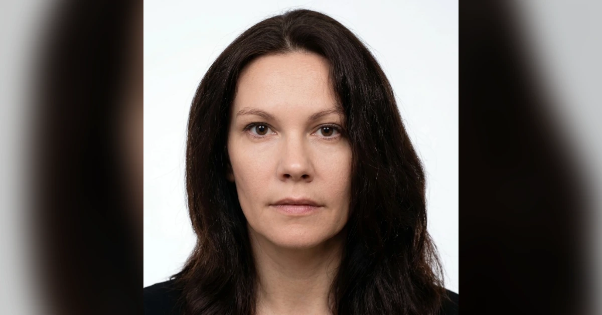 Portrait of a woman with long dark brown hair and a neutral expression against a light background, wearing a black top.