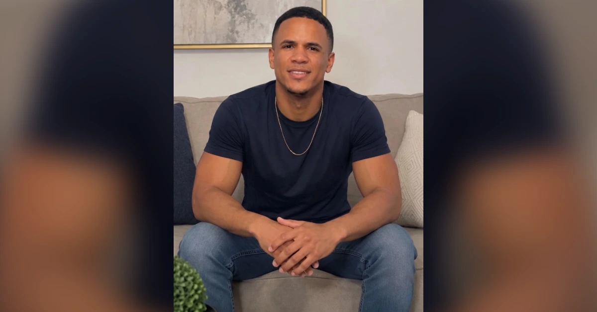 Man wearing a navy T-shirt and jeans sits on a beige sofa with his hands folded in his lap.