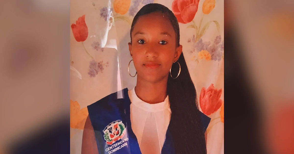 Girl in a white blouse wearing a blue sash with a crest, standing against a floral backdrop.
