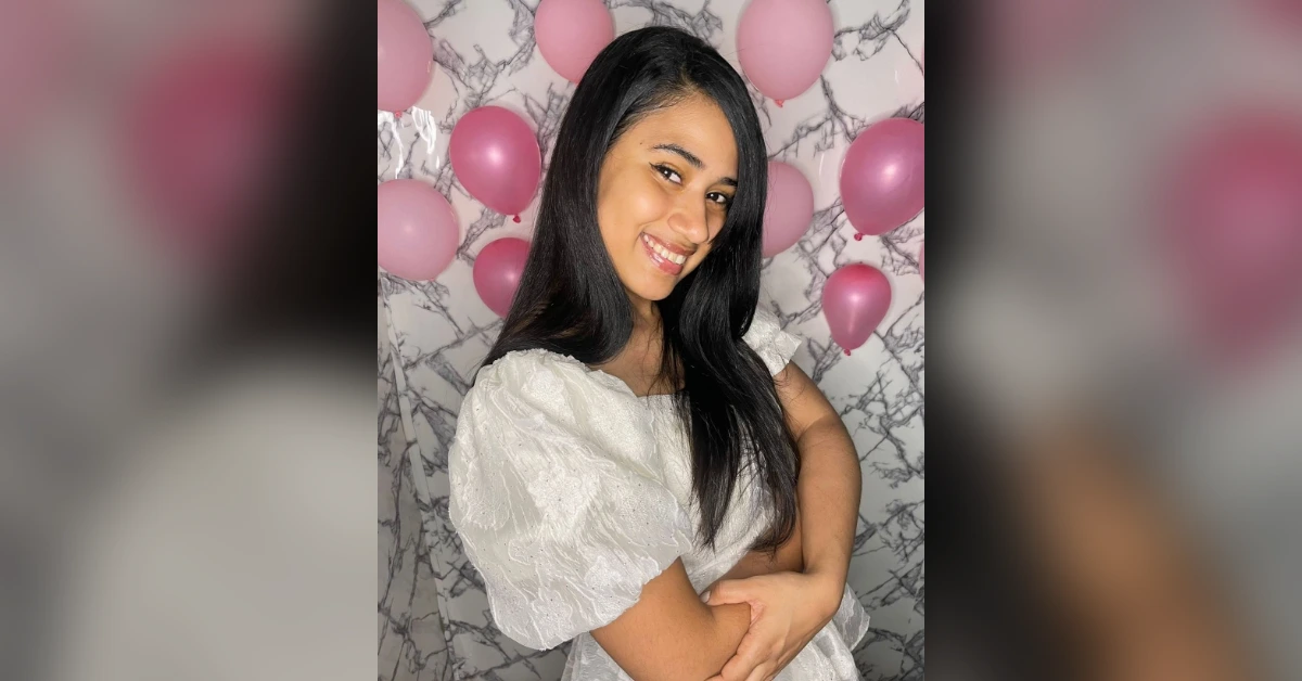 Smiling woman with long dark hair in a white lace dress, arms crossed, pink balloons and a marble backdrop behind her.