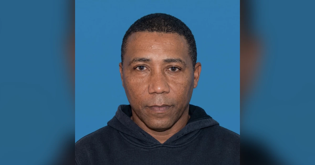 Mugshot-style photo of a man in a dark hoodie against a blue backdrop, facing the camera with a neutral expression.