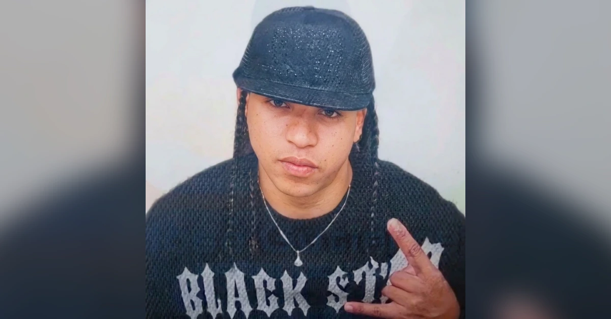 Young person wearing a black sequined cap and braids, looking at the camera, making a rock-on gesture with their right hand.