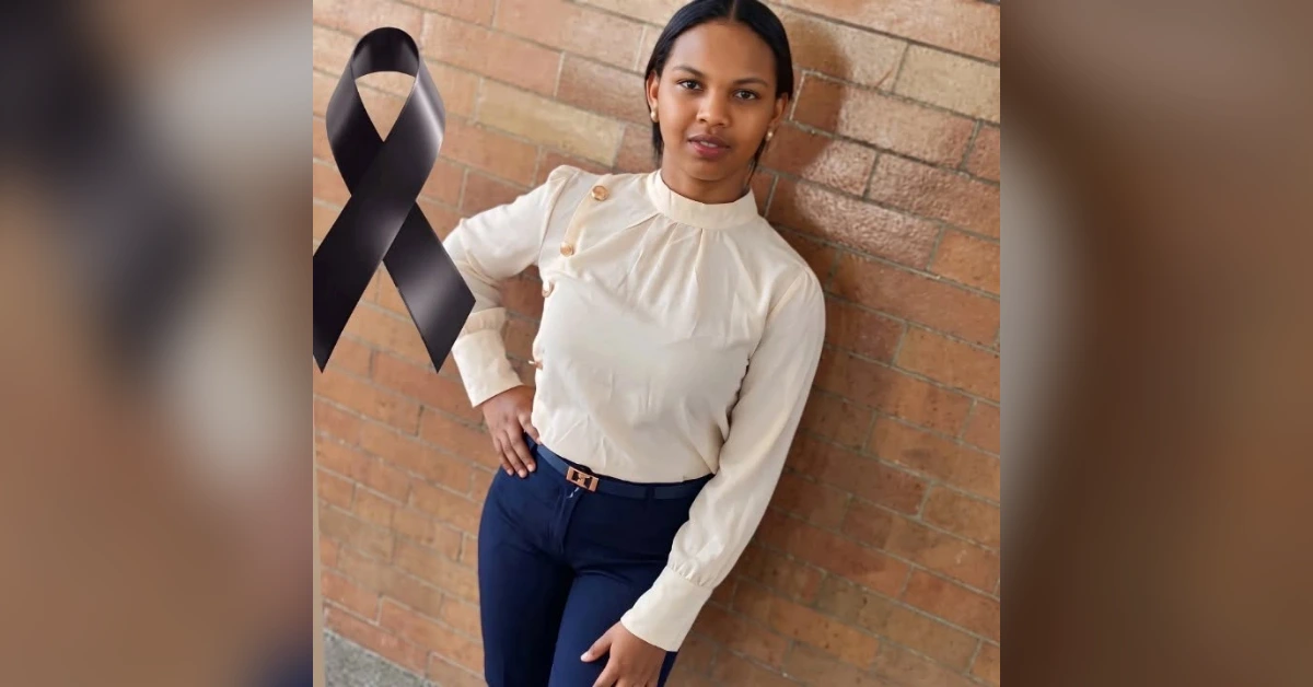 Woman in a cream blouse and navy pants leaning against a brick wall with a large black awareness ribbon beside her, signaling cancer awareness.