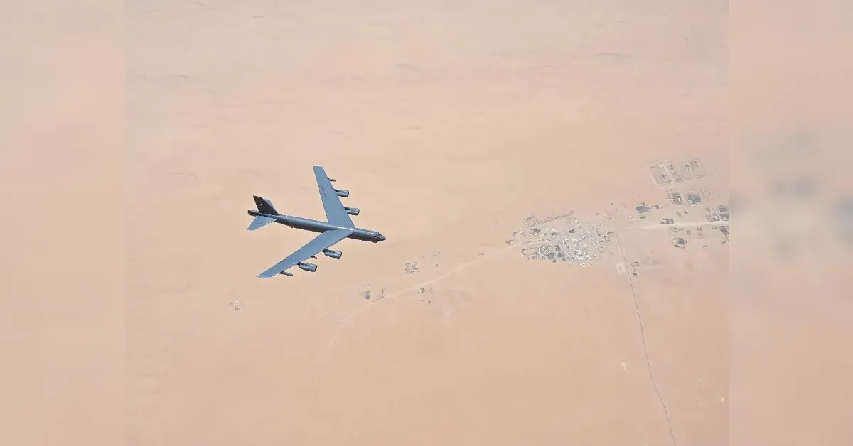 Large military bomber with eight engine pods gliding over a sandy desert with a small village below.