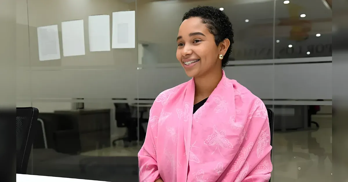 Smiling woman with short curly hair wearing a pink shawl in a glass-walled office setting