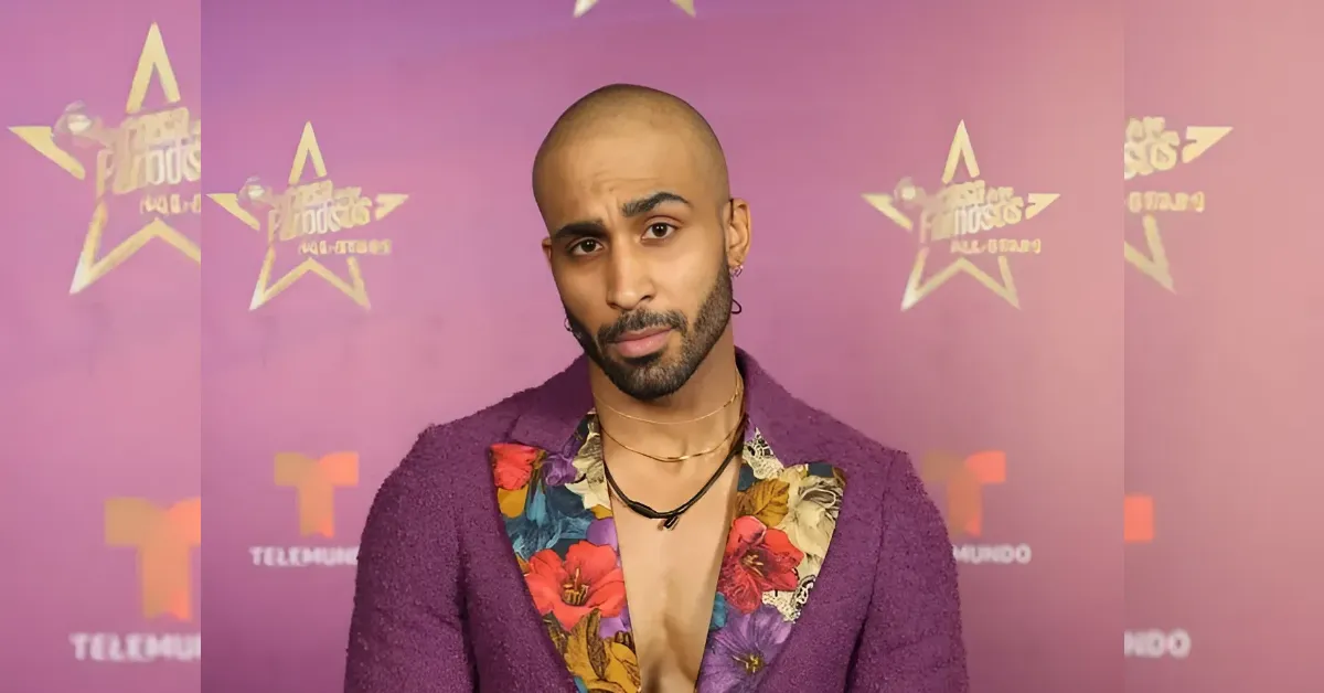 Man in a purple suit with a colorful floral shirt posing on a pink backdrop with star logos; looks to camera with a slight head tilt.