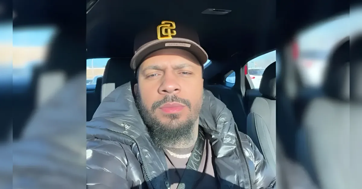 Man taking a selfie inside a car, wearing a dark padded jacket and a black cap with a yellow logo on it.